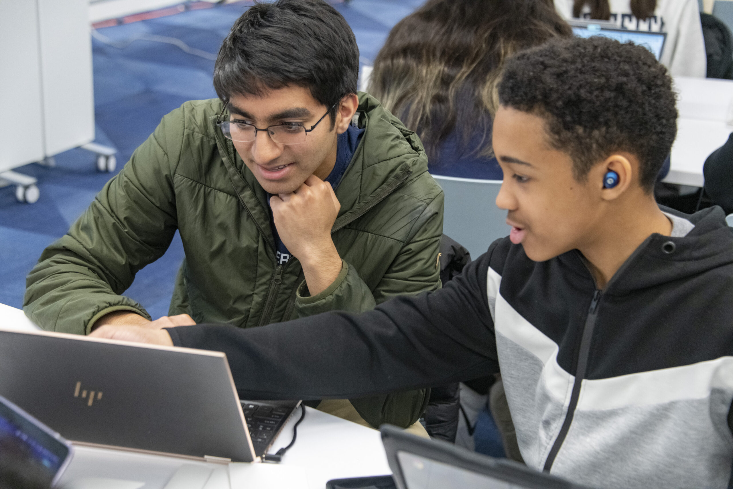 Two students sit next to one another. Both are looking at a laptop, and one is pointing to something on the screen.