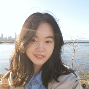 Qingyi has medium length dark hair and dark eyes, she is wearing a tan sport coat over a light blue shirt, facing the camera and smiling.