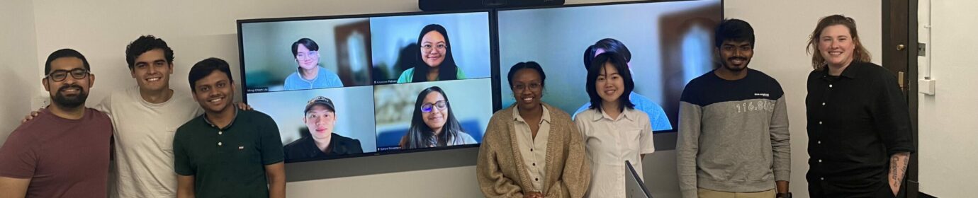 A photo of UChicago graduate student Fellows, some pictured as virtual participants, and Program Manager. Fellows are standing in a row near tables with computers, facing the camera and smiling.